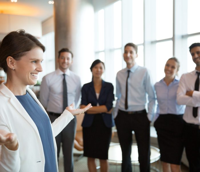 Portrait of smiling businesswoman with open hands gesture standing in office hall, her business team on background
